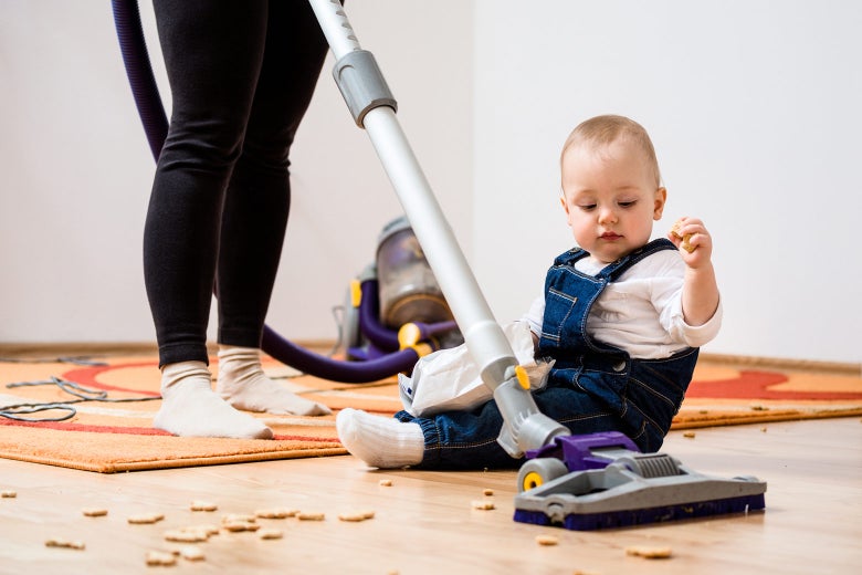 A woman tries to vacuum up crackers as a small child sitting on floor creates more of a mess.